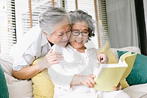 Portrait of beautiful Asian senior couple reading together interesting book while sit on couch in living room at home. Old couple