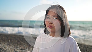Portrait of beautiful Asian teenage girl standing on beach looking away turning to camera smiling. Happy relaxed