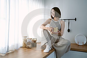 Portrait of beautiful Asian woman in cozy clothes relax sitting on counter top at kitchen drinking coffee and looking through