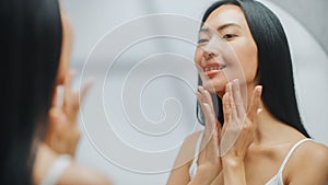 Portrait of Beautiful Asian Woman Gently Applying Face Cream Looking in Bathroom Mirror. Young Adu