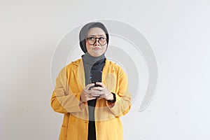portrait of beautiful asian woman in hijab, glasses and wearing yellow blazer thinking while holding mobile phone