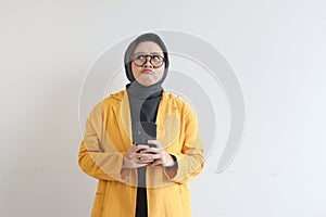 portrait of beautiful asian woman in hijab, glasses and wearing yellow blazer thinking while holding mobile phone