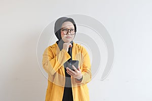 portrait of beautiful asian woman in hijab, glasses and wearing yellow blazer thinking while holding mobile phone