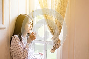 Portrait of beautiful asian woman is holding a cup of coffee and looking something on window at home in the morning,Happy and smil