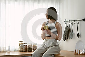 Portrait of beautiful Asian woman holding hot coffee and looking through window while sitting on top counter kitchen. Korean or