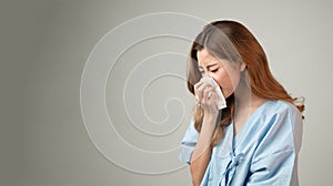 Portrait of a beautiful Asian woman in patient gown hold handkerchief to blow her nose, flu cold sneezing isolated in gray