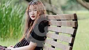 Portrait of beautiful Asian woman sitting on bench in summer forest, Chinese girl in vintage black dress looking at camera