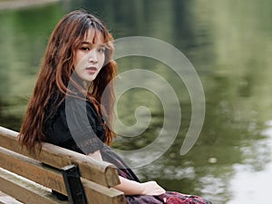 Portrait of beautiful Asian woman sitting on bench in summer forest, Chinese girl in vintage black dress looking at camera