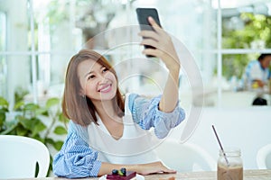 Portrait A beautiful Asian woman in a white shirt sits happily using her cell phone to take a selfie in a bakery