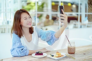 Portrait A beautiful Asian woman in a white shirt sits happily using her cell phone to take a selfie in a bakery