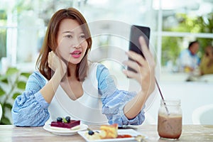 Portrait A beautiful Asian woman in a white shirt sits happily using her cell phone to take a selfie in a bakery