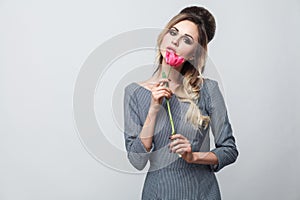 Portrait of beautiful attractive fashion model in grey dress with makeup and hairstyle, standing, holding red tulip and looking at