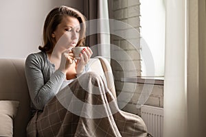 Portrait of beautiful Ñaucasian young woman sitting on sofa covered blanket, drinking hot coffee from cup and looking at window.