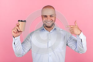 Portrait of a beautiful bald man with a beard dressed in a white shirt. points to a cup of coffee with happy emotions. stands oppo