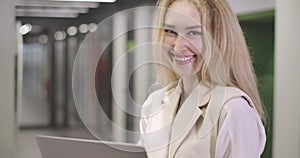 Portrait of beautiful blond Caucasian woman standing with laptop in open space office, looking at camera and smiling