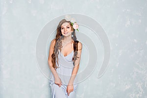 Portrait of a beautiful blond girl with curly hair standing in a studio with a circlet of peonies. She wears light silk