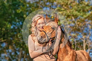 Portrait of a beautiful blond woman with her arms around the neck of her yellow foal. In autumn sunlight, trees in background