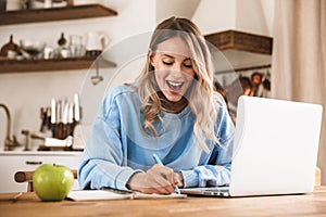 Portrait of beautiful blond woman 20s wearing casual sweatshirt working on laptop and writing down notes at home