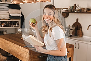 Portrait of beautiful blond woman reading book and drinking tea while standing in stylish wooden kitchen at home