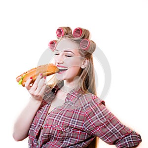 Portrait of beautiful blond young woman in curlers with eyes closed having fun eating hot dog on white copy space background