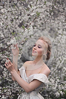 Portrait of a beautiful blonde girl in a luxurious white dress with open shoulders on the background of a blooming orchard.