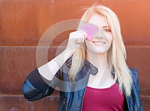 Portrait of a beautiful blonde girl with one hand closes the eyes pink paper heart. Filter, light leak effect.