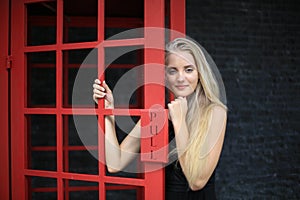 Portrait of Beautiful blonde hair girl on black dress standing in red phone booth against black wall as portrait fashion pose outd
