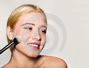 Portrait of beautiful blonde young woman face holding make up brushes. Spa model girl with clean skin isolated on a gray