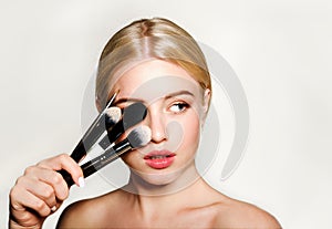 Portrait of beautiful blonde young woman face holding make up brushes. Spa model girl with clean skin isolated on a gray