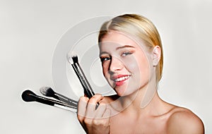 Portrait of beautiful blonde young woman face holding make up brushes. Spa model girl with clean skin isolated on a gray