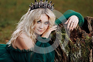 Portrait of A beautiful blonde young woman in a green dress and a diadem on her head in the forest. girl sitting near the old
