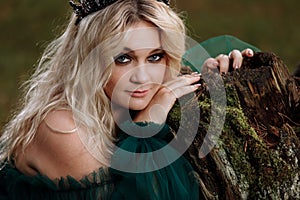 Portrait of A beautiful blonde young woman in a green dress and a diadem on her head in the forest. girl sitting near the old