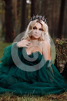 Portrait of A beautiful blonde young woman in a green dress and a diadem on her head in the forest. girl sitting near the old