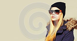 Portrait of beautiful blonde young woman looking away wearing hat, sunglasses and jacket on gray background, blank copy space for