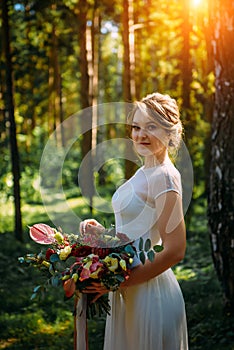 Portrait of a beautiful bride among green trees on sunny summer day. Young girl in a white wedding dress
