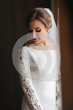 Portrait of beautiful bride standing by the window at home. Charming bride in white wedding dress