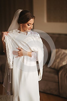 Portrait of beautiful bride standing by the window at home and trying on wedding dress. Charming bride with white