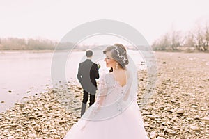 Portrait of beautiful bride wearing white dress and veil with groom standing in background near river