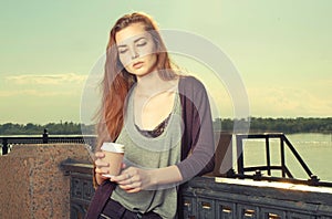 Portrait of beautiful brown haired teen girl standing and looking down. She keeping takeaway drink. Urban city scene