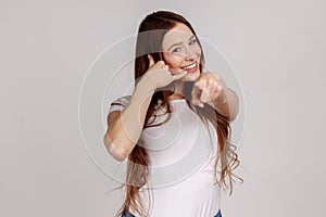 Portrait of beautiful brown haired woman standing with call gesture, looking and pointing at camera.