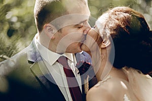 Portrait of beautiful brunette bride kissing handsome groom with red flower boutonniere face close-up, newlywed couple posing