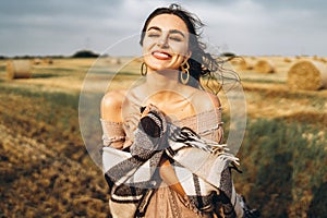 Portrait of a beautiful brunette in a dress and with a warm plaid. Woman enjoying a walk in a wheat field with hay bales
