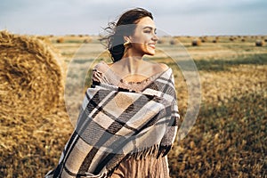 Portrait of a beautiful brunette in a dress and with a warm plaid. Woman enjoying a walk in a wheat field with hay bales