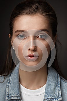 Portrait of beautiful brunette girl with hair tied back dressed in jeans jacket on the dark background in the studio