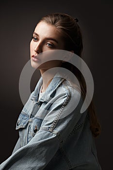 Portrait of beautiful brunette girl with hair tied back dressed in jeans jacket on the dark background in the studio