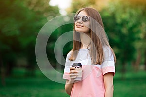 Portrait of beautiful brunette girl in sunglasses walking down the street. Keeping takeaway drink in hand