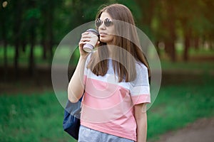 Portrait of beautiful brunette girl in sunglasses walking down the street. Keeping takeaway drink in hand. Smiling.