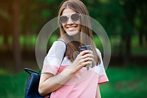 Portrait of beautiful brunette girl in sunglasses walking down the street. Keeping takeaway drink in hand. Smiling.