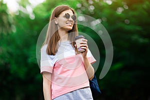 Portrait of beautiful brunette girl in sunglasses walking down the street. Keeping takeaway drink in hand. Smiling