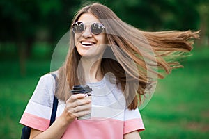 Portrait of beautiful brunette girl in sunglasses walking down the street. Keeping takeaway drink in hand. Smiling. Urban city